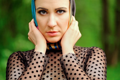 Crop serious female wearing silk headscarf holding face in hands and looking at camera attentively while standing in lush garden
