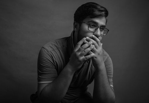 Free Black and white portrait of a man with glasses sitting pensively in a studio setting. Stock Photo