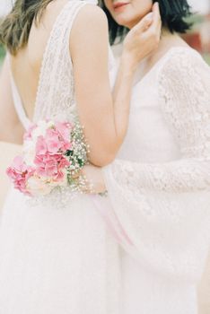 A tender moment between a lesbian couple in elegant wedding gowns with a pink floral bouquet.