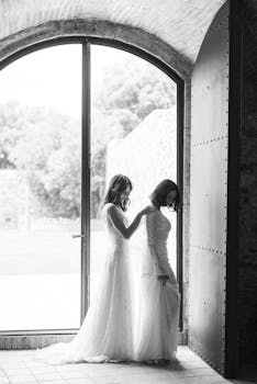 Intimate black and white photo of a same-sex couple in wedding dresses by a large doorway.