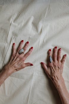 特写镜头中，老年人双手戴着戒指，红色指甲油在织物表面闪闪发光