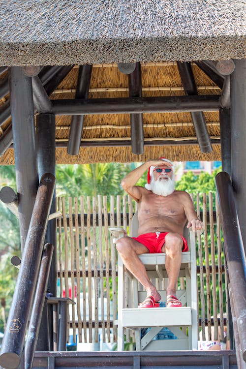 Topless Man in Red Shorts Sitting on Wooden Chair While Looking Out