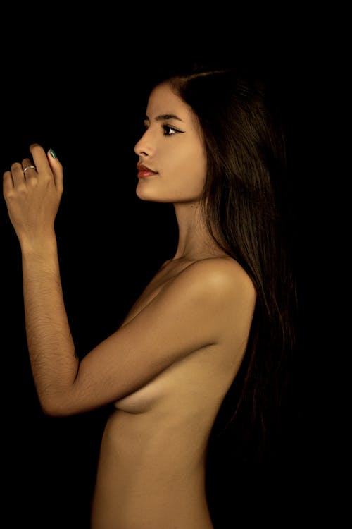 Side view of gorgeous nude female showing perfect body while covering bare chest against black background
