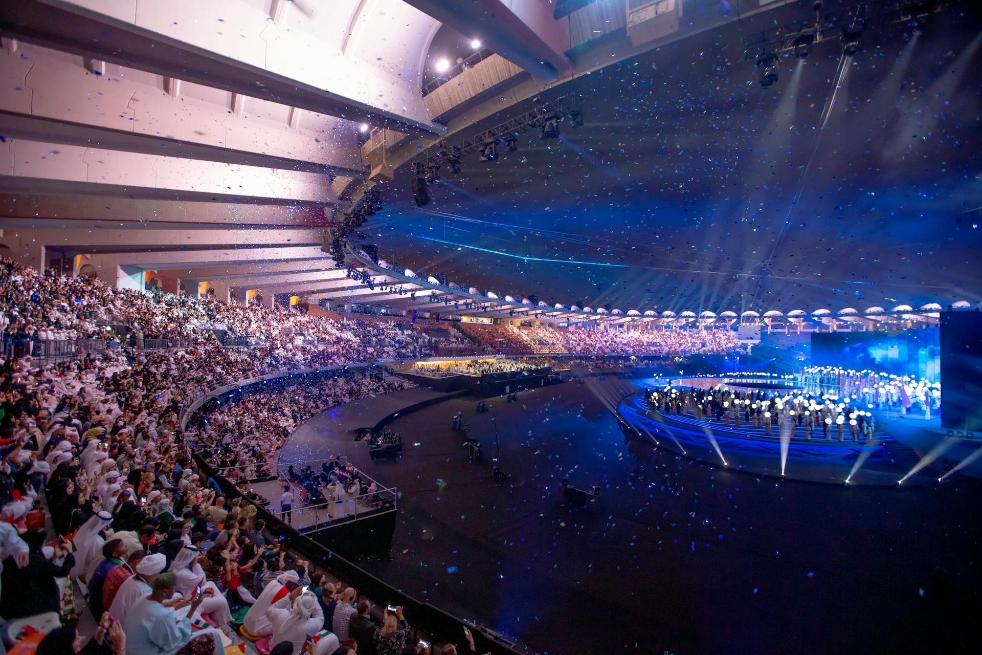 Massive arena concert with vibrant lights, packed audience, and dynamic stage performance.