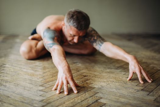 A shirtless man with tattoos stretches on an indoor wooden floor, showcasing wellness and flexibility.
