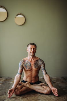 Tattooed man sitting in lotus pose, practicing yoga meditation indoors.