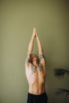 Tattooed man in a yoga pose with closed eyes indoors against a muted green backdrop.