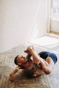 A tattooed man performs a complex yoga pose in a sunlit room, highlighting fitness and flexibility.