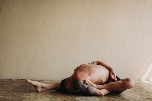 Tattooed man practicing a yoga pose indoors on a wooden floor, showcasing flexibility and strength.