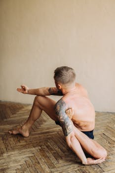 A tattooed man in a yoga pose indoors, highlighting flexibility and focus.
