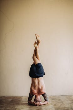 A tattooed man performs a headstand yoga pose indoors, demonstrating strength and balance.