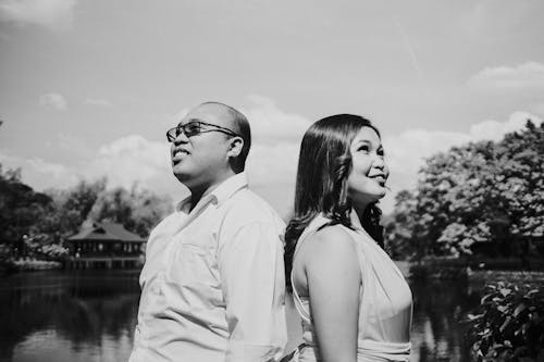 Black and white side view of cheerful ethnic couple admiring cloudy sky while looking up against pond and trees in sunlight