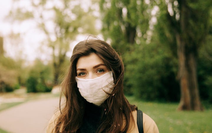 A young woman with a face mask stands outdoors in a park with tall trees, signifying pandemic precautions.
