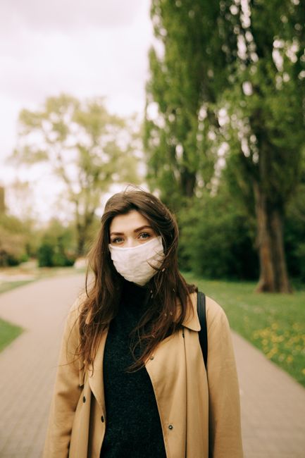 A young woman with a face mask stands outdoors in a park with tall trees, signifying pandemic precautions.