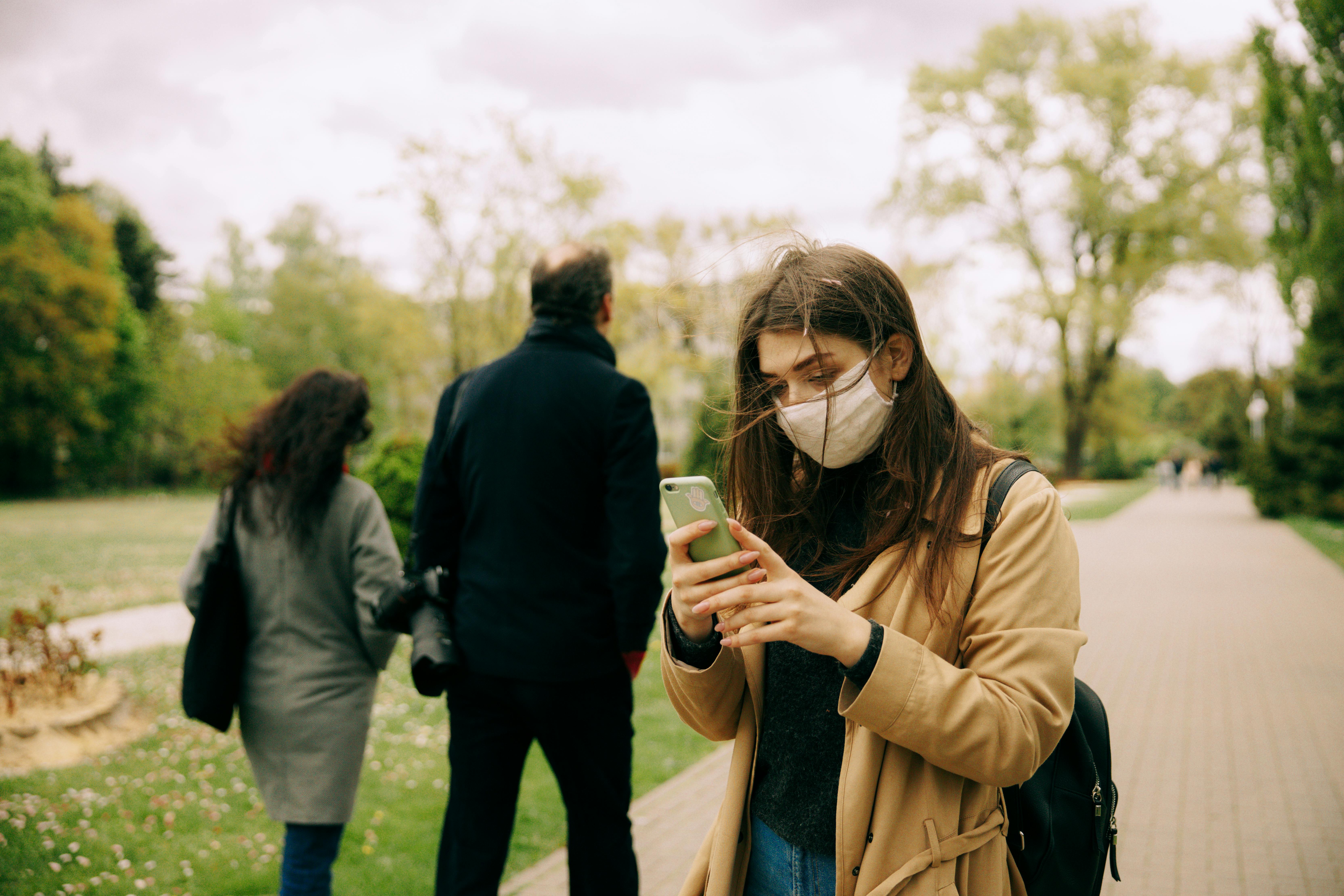 A woman wearing a mask uses her phone in a scenic park setting.