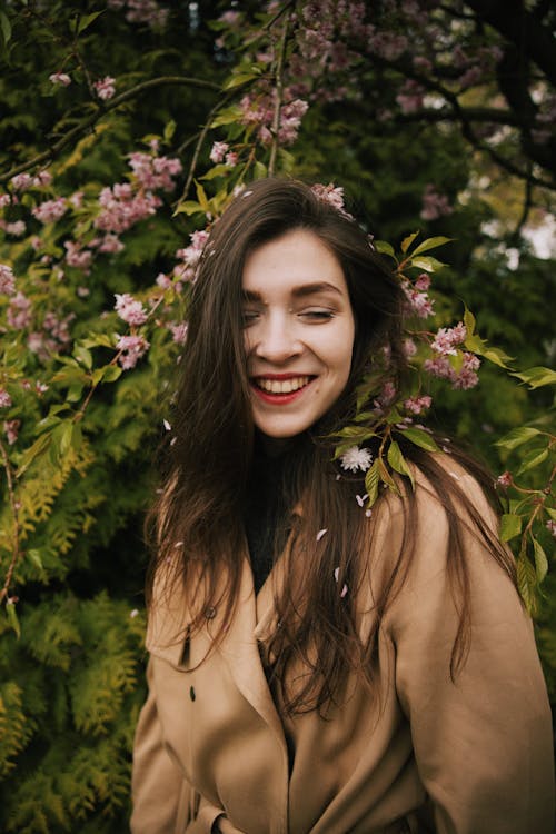 Photo of Woman in Brown Coat Smiling While Standing Near Pink Flowers