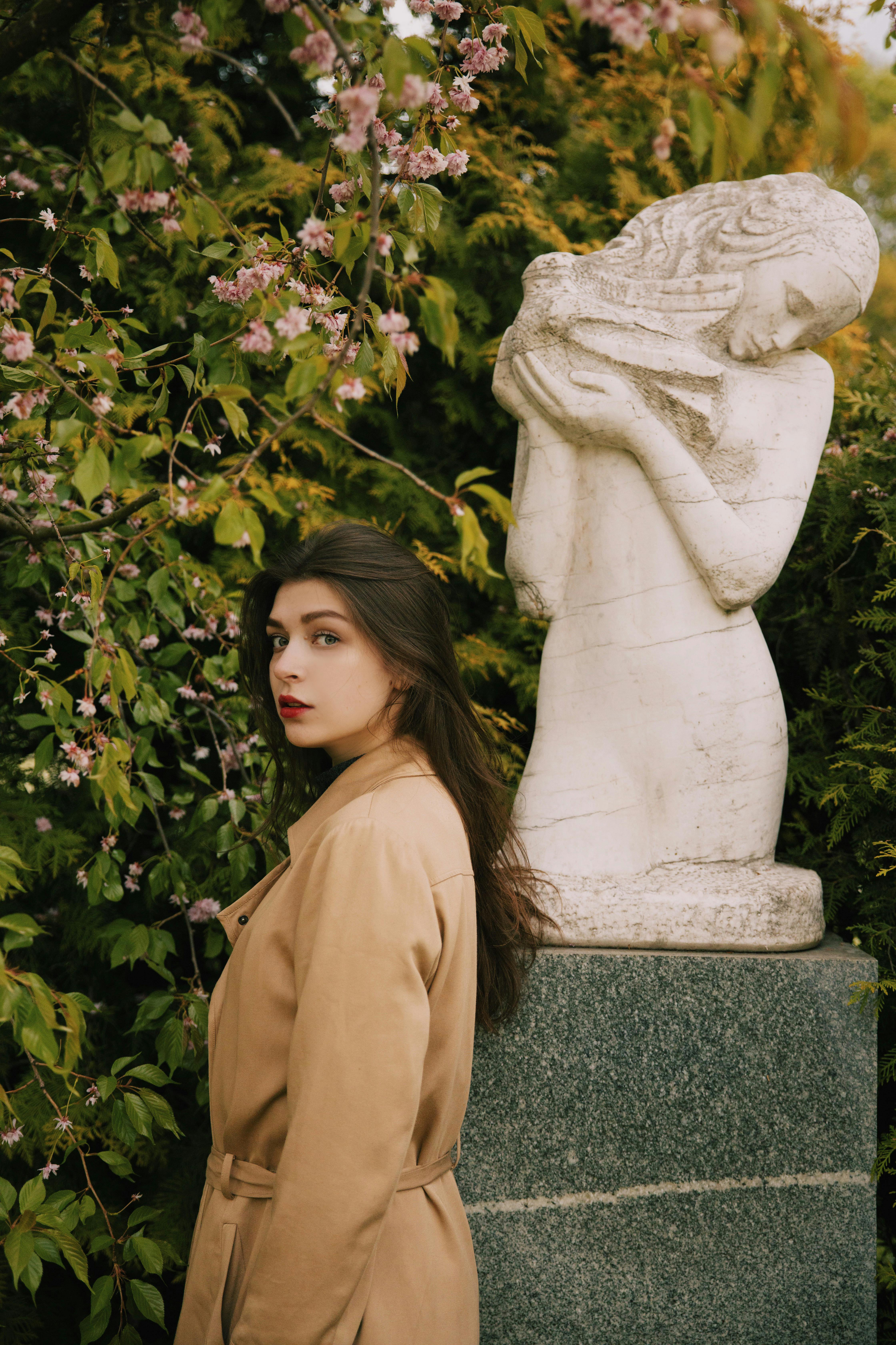 穿着棕色外套的女人站在花园雕像旁边，周围环绕着粉红色的花朵