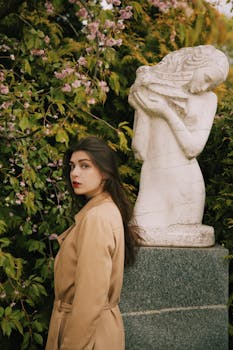 一位穿着棕色外套的女人站在一座花园雕像旁，雕像周围环绕着粉色花朵