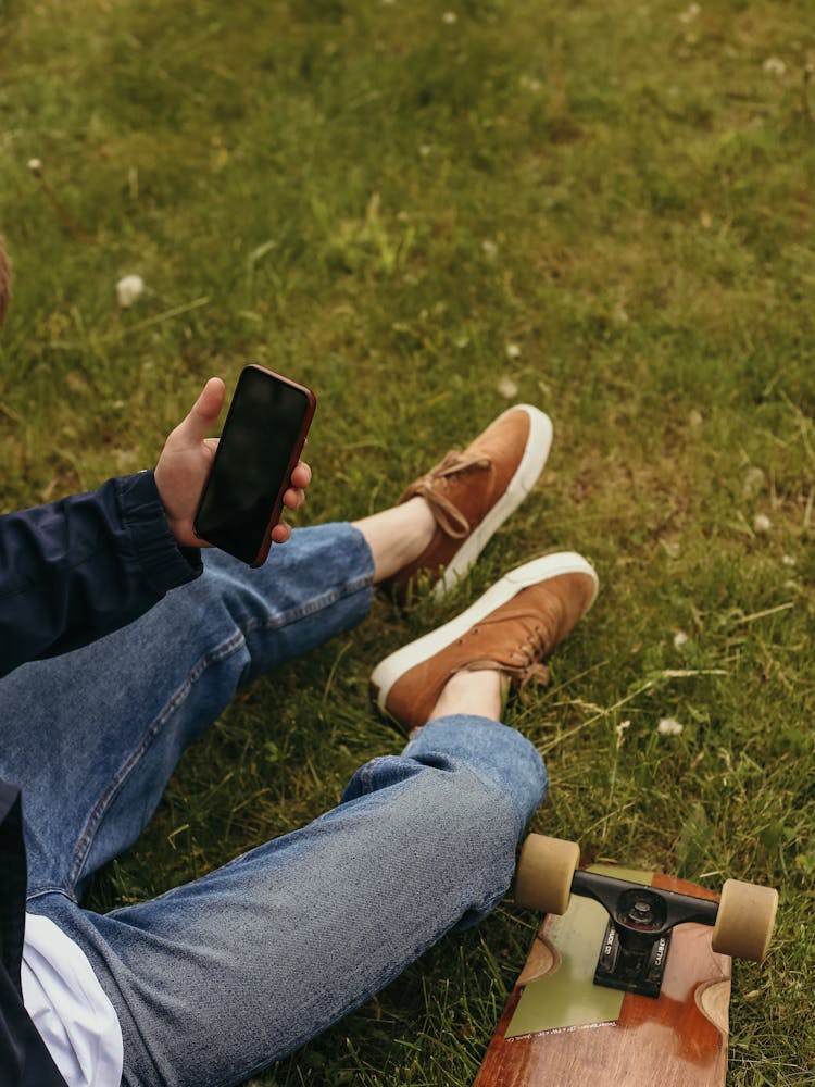 A Person Sitting On The Green Grass Beside The Skateboard Using A Phone