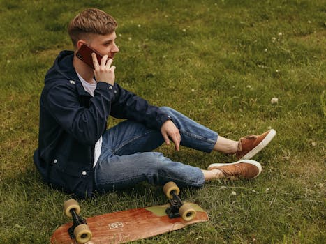 Casual young man talking on the phone while sitting on grass with skateboard, enjoying a sunny day.