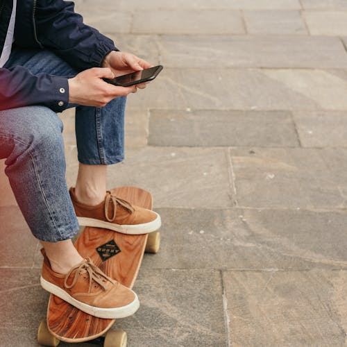 Person in Blue Denim Jeans and Brown Shoes Using Smartphone