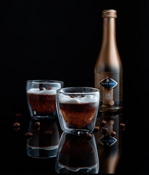 Stylish shot of delicious coffee liqueur with whipped cream and bottled liquor on reflective black surface.
