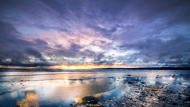 Stunning sunset with dramatic clouds over a calm ocean shore, reflecting colors on the wet sand.