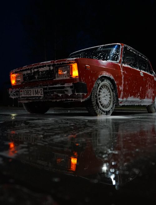 Free Vintage red car being washed at night, reflecting on wet pavement with lights on. Stock Photo