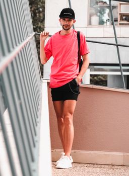Stylish male model in pink shirt and cap showcasing urban fashion near metal railing.