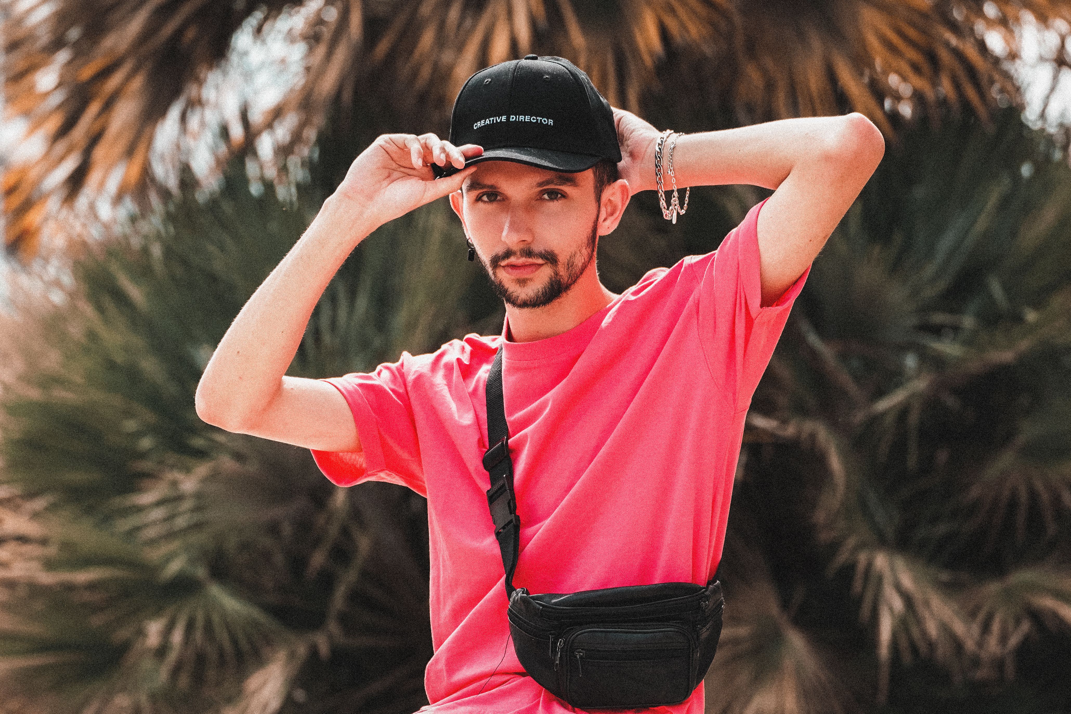 Fashionable man in pink shirt adjusts cap in a stylish pose outdoors, showcasing street fashion.