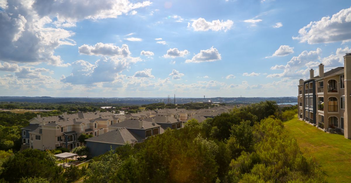 Modern multi storey buildings surrounded by tall verdant trees in park under light blue sky with clouds