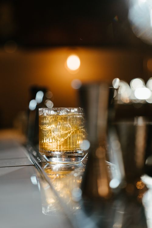 Free A glass of whiskey with ice on a reflective bar surface, surrounded by warm ambient lighting. Stock Photo