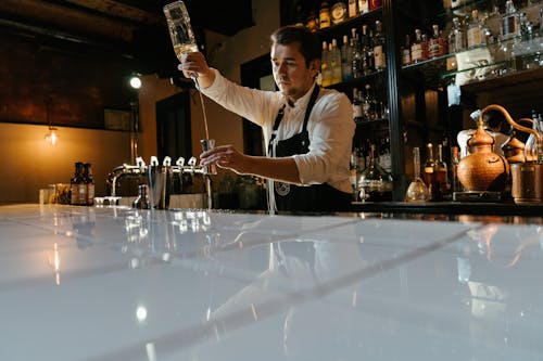 Man in White Long Sleeve Shirt Holding Bottle Pouring Water on Clear Drinking Glass