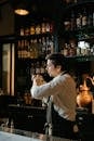 Man in White Long Sleeve Shirt Pouring Liquor on Clear Glass Bottle