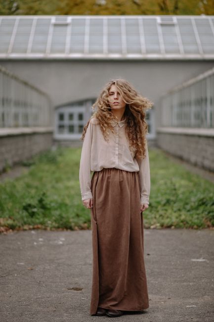 A woman with curly hair stands outdoors wearing a long brown skirt and blouse.