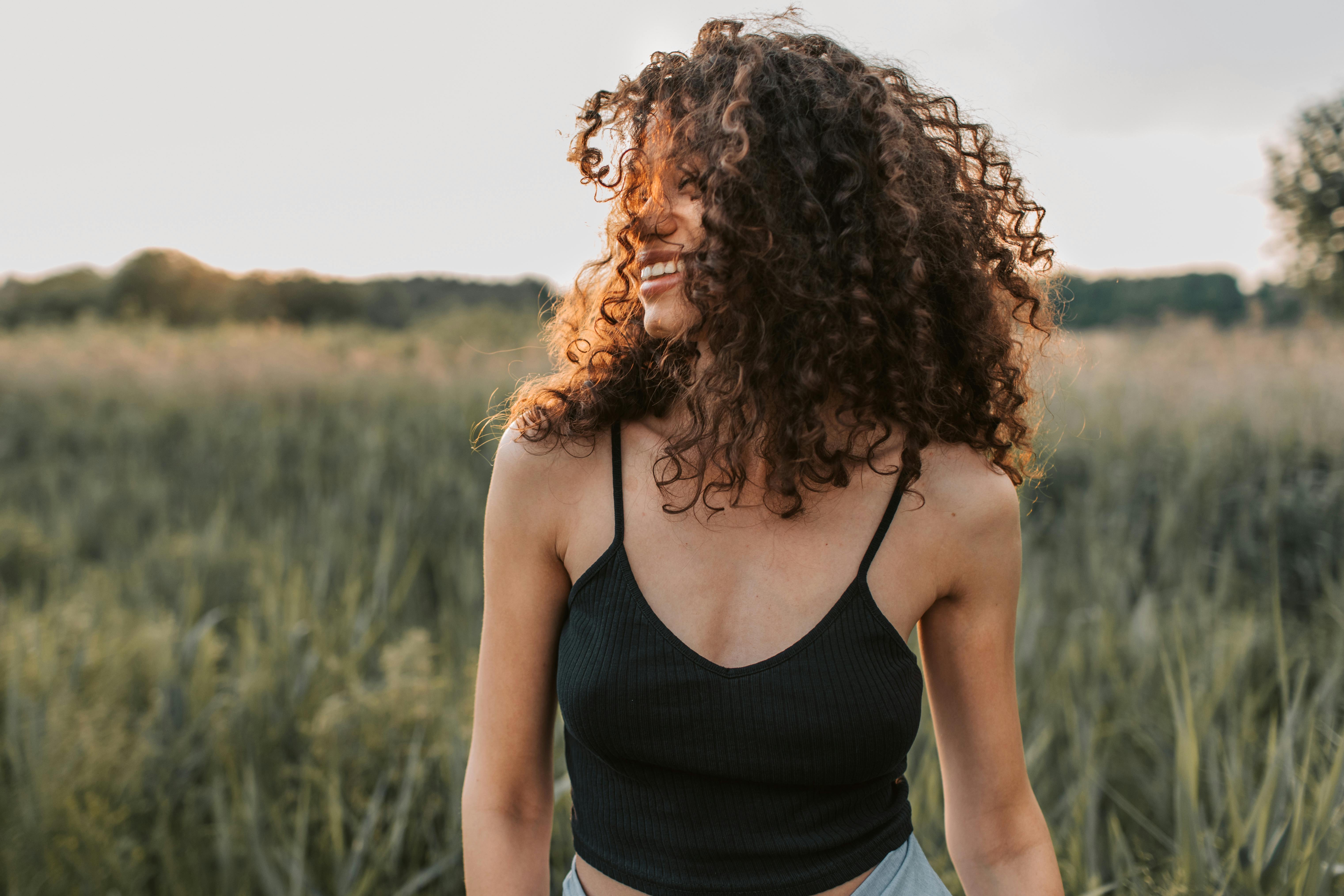 A smiling woman with curly hair stands in a grassy field, wearing a black tank top under soft, golden sunlight.