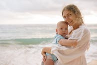 母亲抱着婴儿，慈爱地站在海滩上，脸上洋溢着快乐的微笑，身后是大海