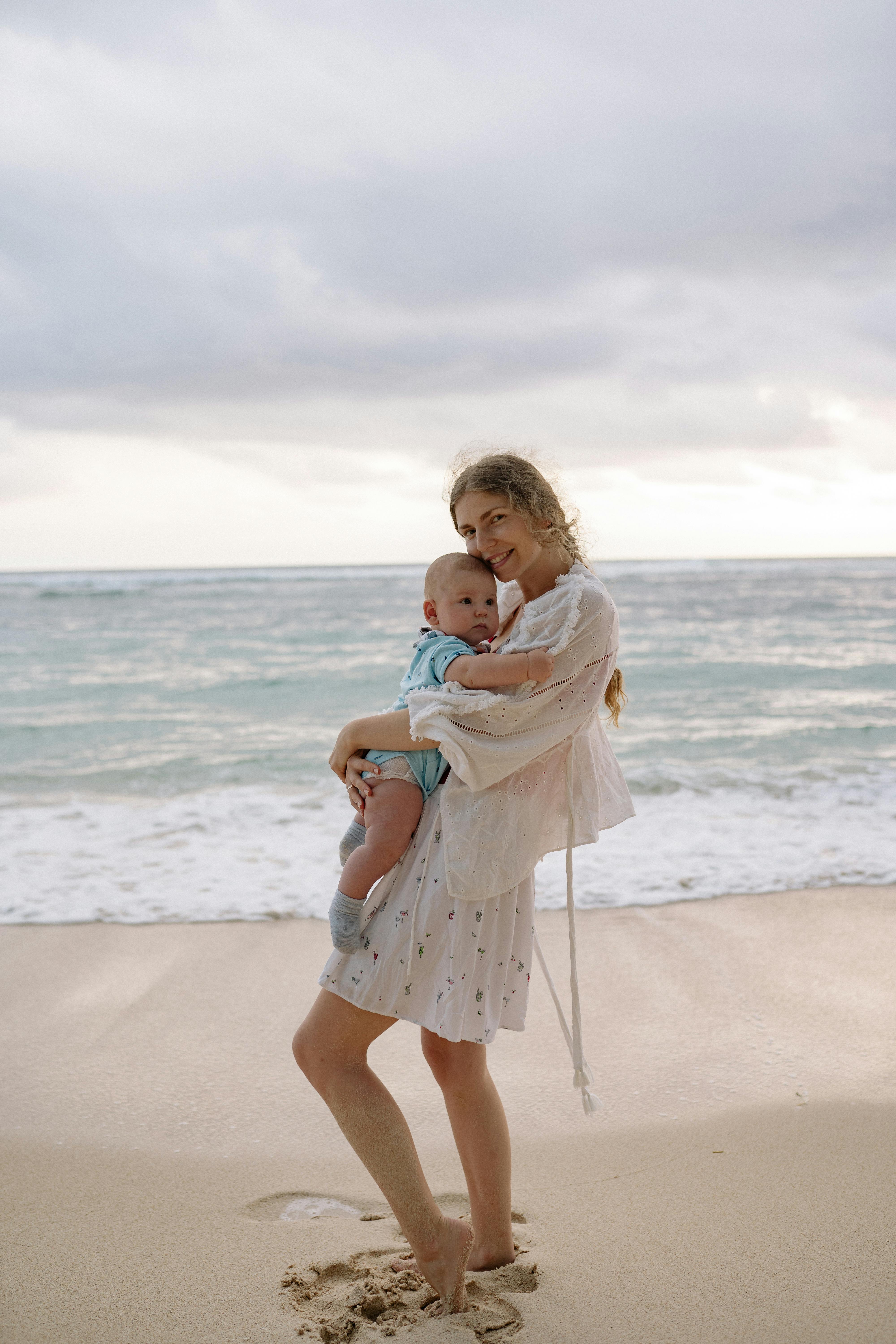 一位母亲抱着她的孩子，在阴沉天空下的沙滩上，海浪轻柔