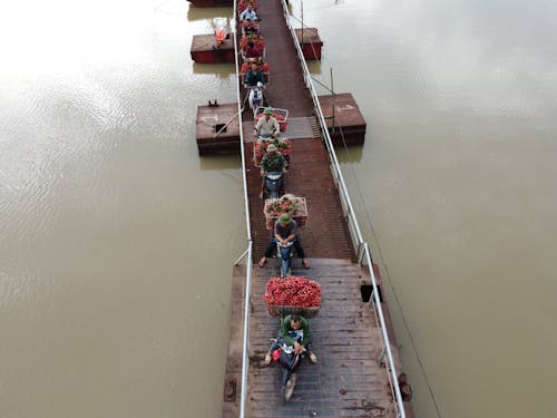 From above of people transporting food in baskets on motorbikes riding on float bridge above rippled river