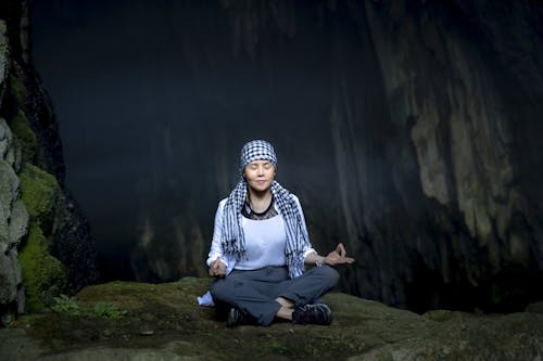 Full body of calm female with closed eyes in butterfly pose with okay gesture in rocky cave