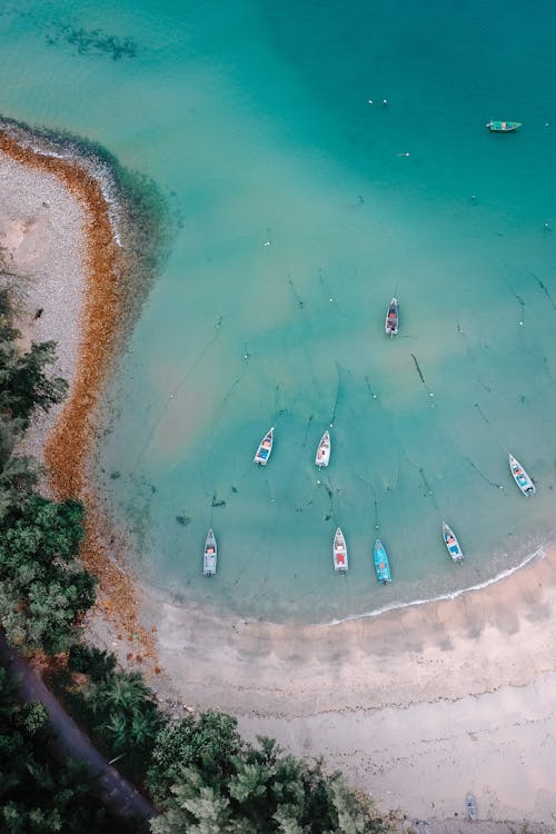 Breathtaking aerial view of boats in turquoise water of wavy ocean washing sandy beach surrounded by green trees