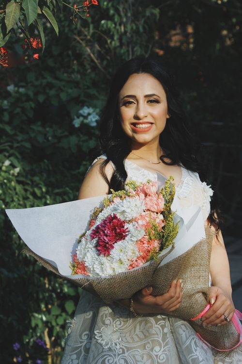 Free Beautiful smiling ethnic female in elegant white dress holding fragrant bouquet while standing in summer garden and looking at camera Stock Photo