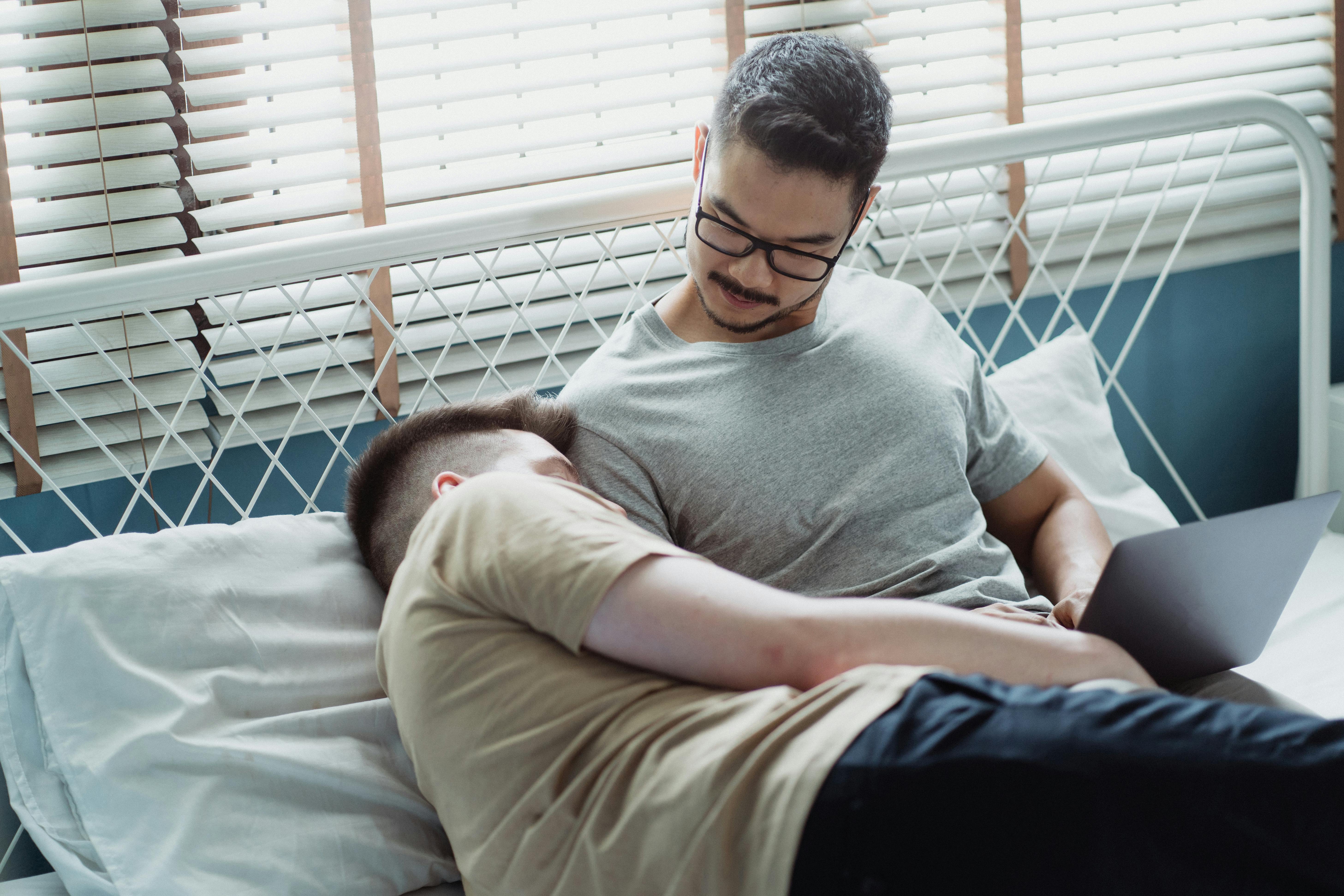 man cuddling against another man using a laptop in bed