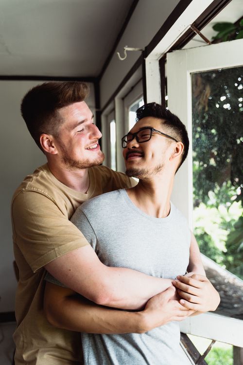 Free Smiling interracial gay couple in a warm embrace by the window, showcasing love and happiness. Stock Photo