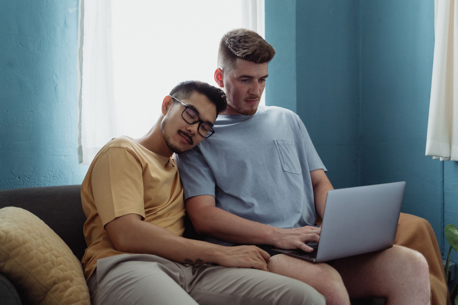 A loving interracial gay couple relaxes indoors, one asleep on the other's shoulder, while using a laptop.