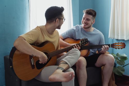 Free Two men sitting together, playing guitar and smiling in a cozy living room. Stock Photo
