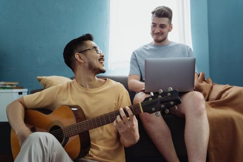 Free Two friends relax together, one playing guitar and the other using a laptop, creating a joyful atmosphere indoors. Stock Photo