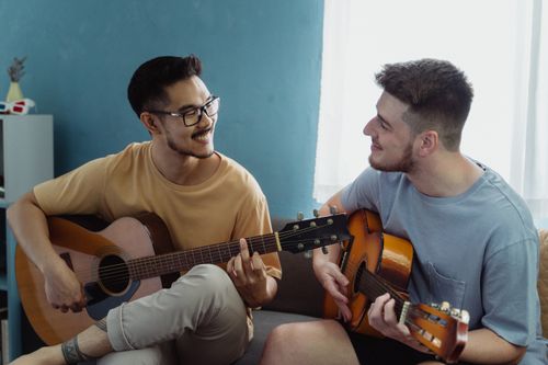 Free Two young men playing guitars and smiling in a cozy living room setting, enjoying music together. Stock Photo