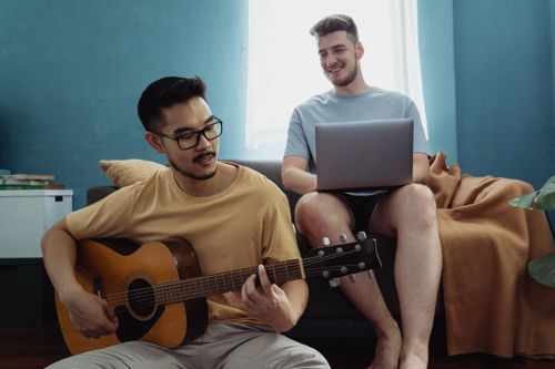 Free Casual scene of two friends in a living room, one playing guitar, the other using a laptop. Stock Photo