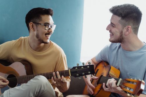 Free Two friends sitting in a living room, playing acoustic guitars and smiling with joy. Stock Photo